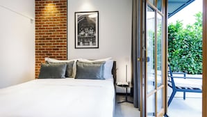 A modern bedroom with a brick accent wall, a large bed with grey pillows, a framed picture above the bed, and a view of greenery through a sliding glass door. - OLD TOWN SNAIL Design Apartments (Bucharest)