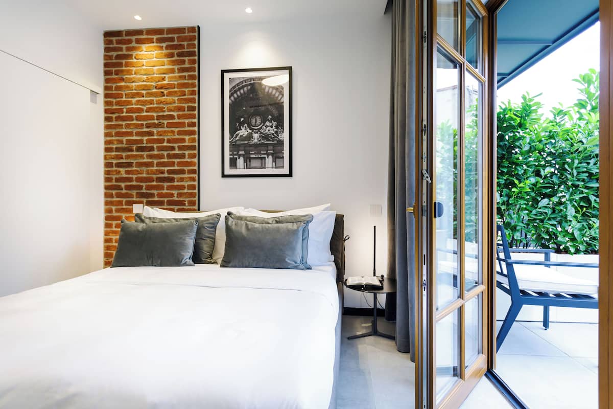 Un dormitorio moderno con una pared de ladrillo decorativa, una cama grande con almohadas grises, un cuadro enmarcado sobre la cama y vistas a la vegetación a través de una puerta corrediza de vidrio.