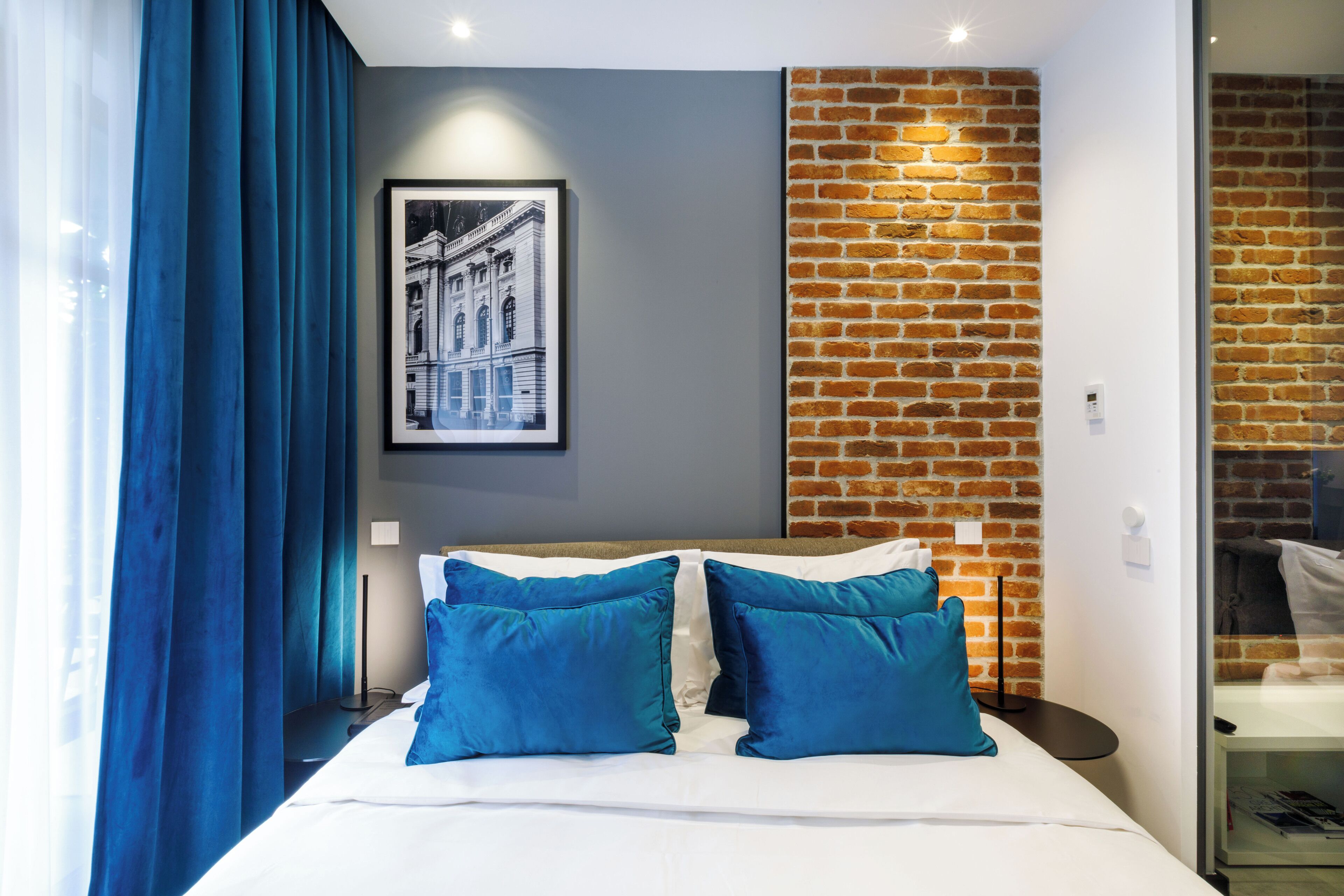 Quarto de hotel moderno com uma cama grande, almofadas azuis, um quadro na parede e uma parede de tijolos decorativa.