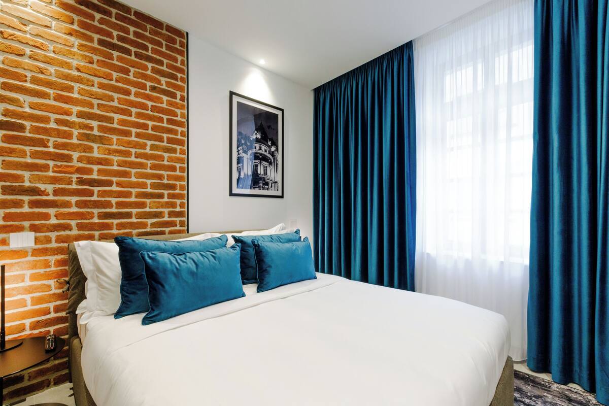 Una habitación de hotel con una pared de ladrillo, una cama grande con ropa blanca y almohadas azules, un cuadro en la pared y una ventana con cortinas azules.
