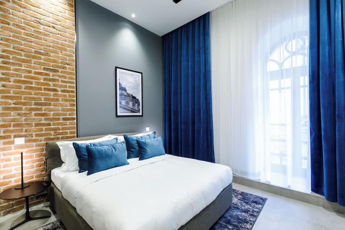 Una habitación de hotel moderna con una pared de ladrillo decorativa, una cama grande con ropa de cama azul y blanca, un cuadro en la pared y vistas a través de cortinas traslúcidas.