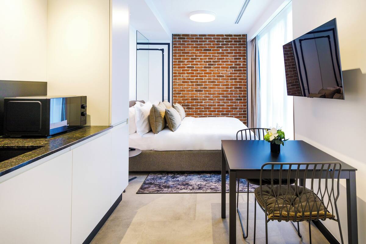 Una habitación de hotel compacta con kitchenette, una cama con almohadas, una mesa de comedor con sillas y una pared de ladrillo decorativa.