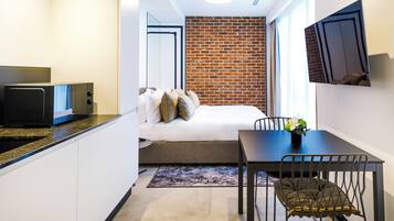 Una habitación de hotel compacta con kitchenette, una cama con almohadas, una mesa de comedor con sillas y una pared de ladrillo decorativa.