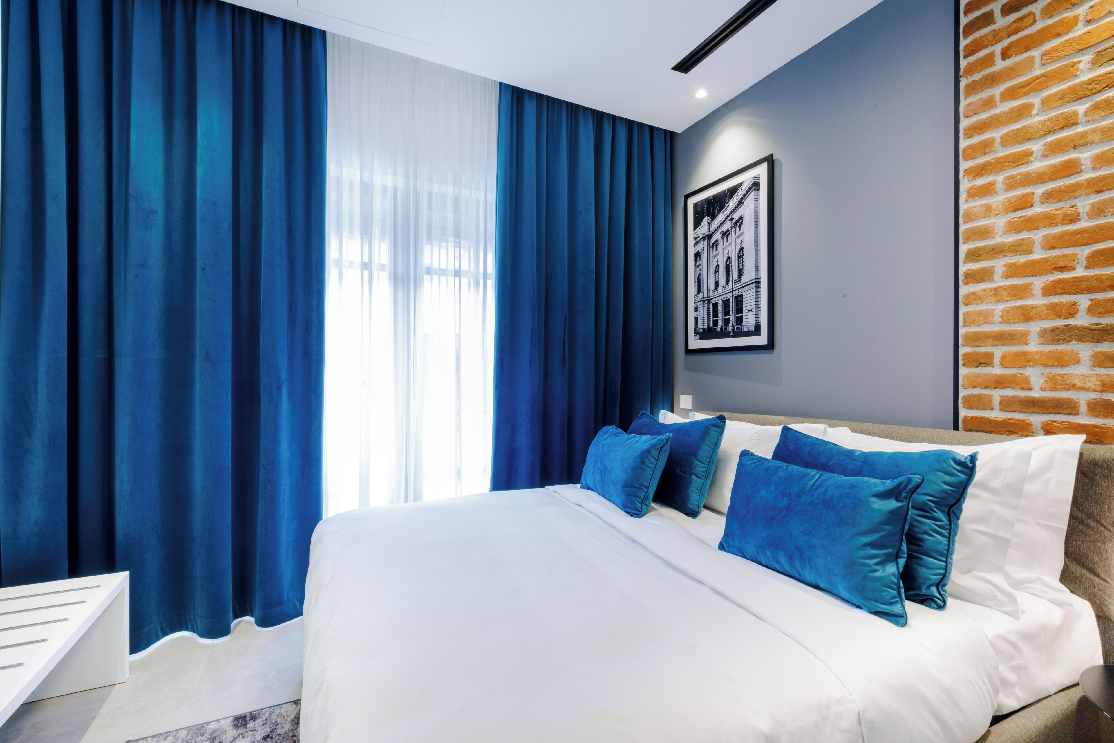 A modern hotel room with a large bed, blue curtains, a brick accent wall, and a framed picture on the wall.