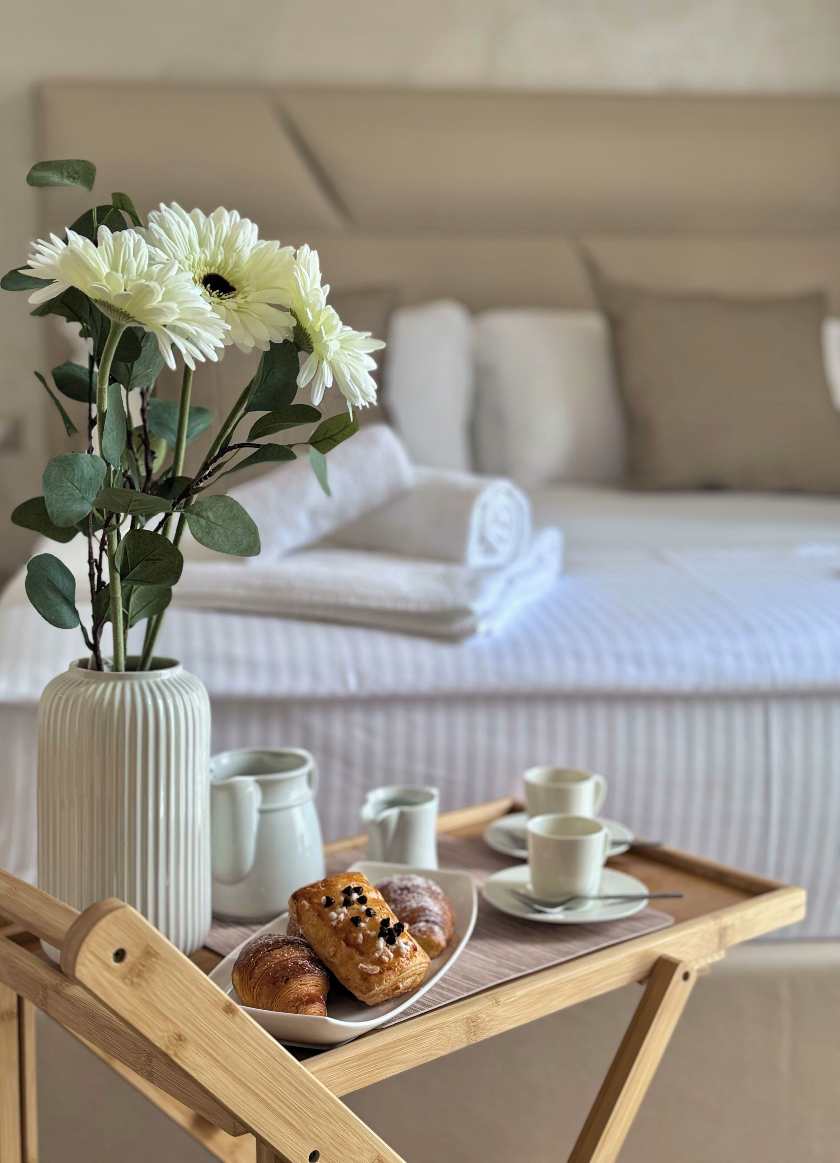 Ein ordentlich bezogenes Bett mit Kissen und einer zusammengefalteten Decke, eine Vase mit weißen Blumen, ein Tablett mit Gebäck und Tassen sowie ein Holzhocker.
