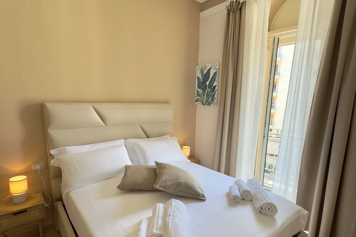 Una cama bien hecha con almohadas y toallas enrolladas, una lámpara de noche y vistas al exterior a través de una ventana con cortinas.
