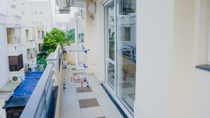 A narrow corridor with a tiled floor, a metal handrail, and a glass door leading to a balcony with a view of neighboring buildings and a tree. - Lime Tree Hotel Golf Course Extension Rd (Gurugram)