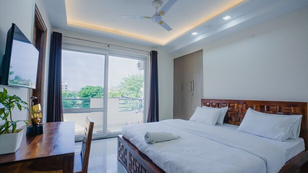 A hotel room with a bed, a desk, a chair, a ceiling fan, and a view of the outdoors. - Lime Tree Hotel Golf Course Extension Rd (Gurugram)