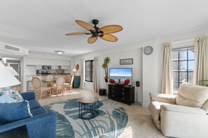 Una sala moderna con ventilador de techo, televisor, sofá, mesa de centro y vista al océano a través de una ventana.