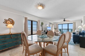 Una sala de estar moderna con una mesa de comedor de vidrio, asientos cómodos, un ventanal con vistas y un ventilador de techo.
