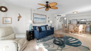 Una sala de estar con un sofá azul, un sillón blanco, un ventilador de techo, un comedor con sillas de madera y una cocina al fondo.