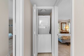 Une pièce compacte équipée d’un lave-linge et d’un sèche-linge intégrés, d’un lit recouvert d’une couvre-lit à motifs floraux et d’une petite table de chevet avec une lampe.