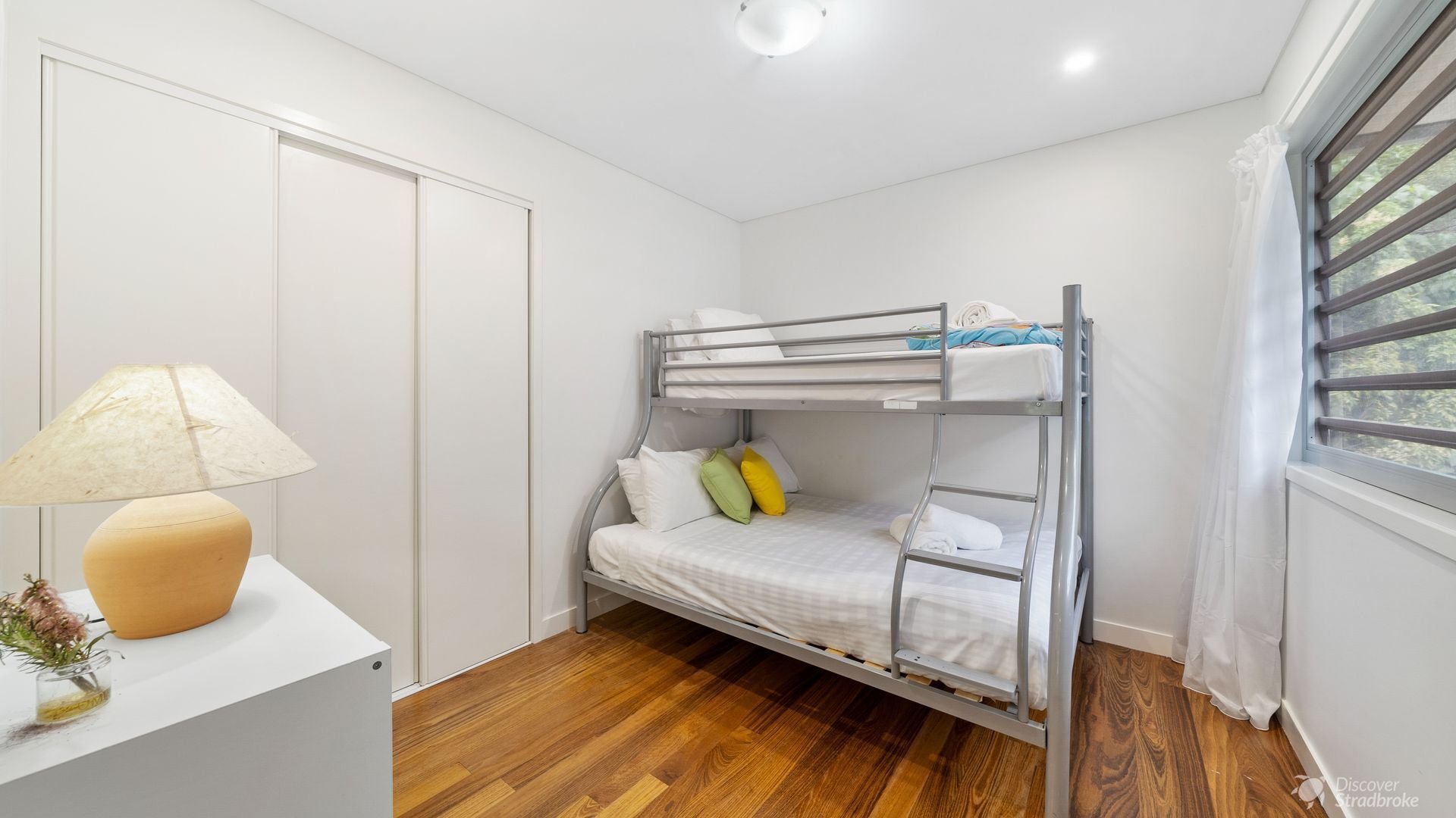 A bunk bed room with a wooden floor, a white desk with a lamp, and a window with curtains.