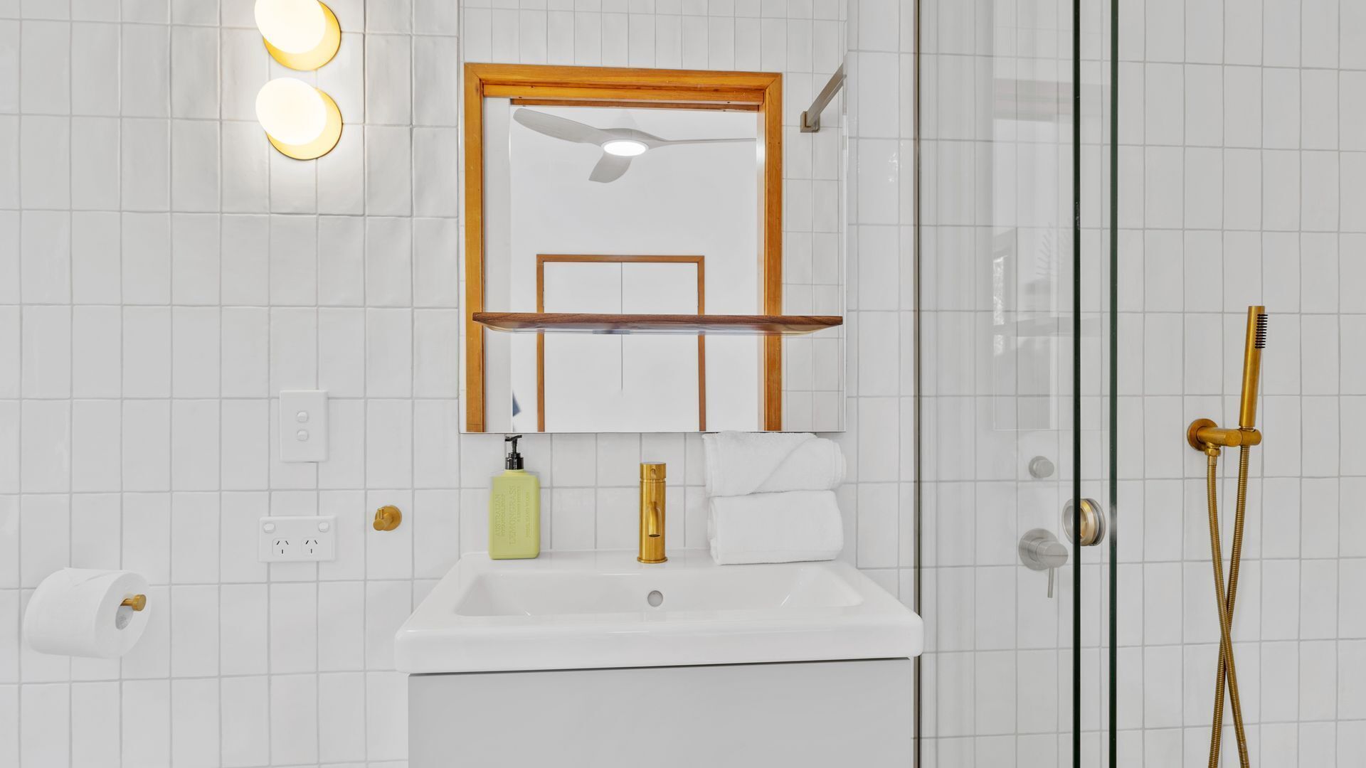 Un baño con un lavamanos blanco, un espejo con marco de madera, un perchero para toallas y una ducha con detalles dorados.