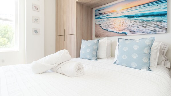 A neatly made bed with white linens and blue pillows with shell patterns, a wooden wardrobe, and a wall painting of a beach scene.