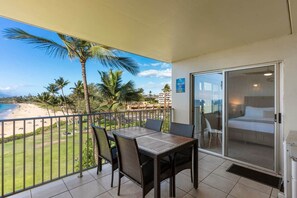 A balcony with a view of the beach, a dining table with chairs, and a bedroom with a bed and a sliding door.