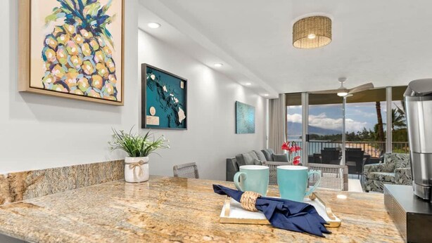 A modern kitchen with granite countertops, a coffee maker, and a dining area with a view. - Royal Mauian 503-Oceanfront with AC (Kihei)