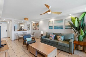 A modern living room with a sofa, coffee table, and chairs. There is a ceiling fan, artwork on the walls, and a large plant.