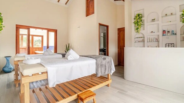 A spa room with a wooden massage table, a small wooden stool, and a wall-mounted shelf with toiletries. - Villa Santa Ana Premium (Villa de Leyva)