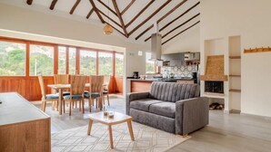A spacious living area with a sofa, dining table, and chairs, a kitchen in the background, and large windows offering a view of the outdoors. - Villa Santa Ana Premium (Villa de Leyva)