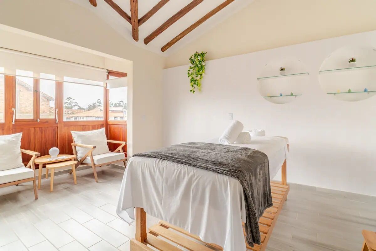 A bedroom with a bed, a wooden bench, a chair, a small table, a window, and a wall-mounted shelf with plants.