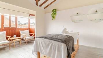 A bedroom with a bed, a wooden bench, a chair, a small table, a window, and a wall-mounted shelf with plants.