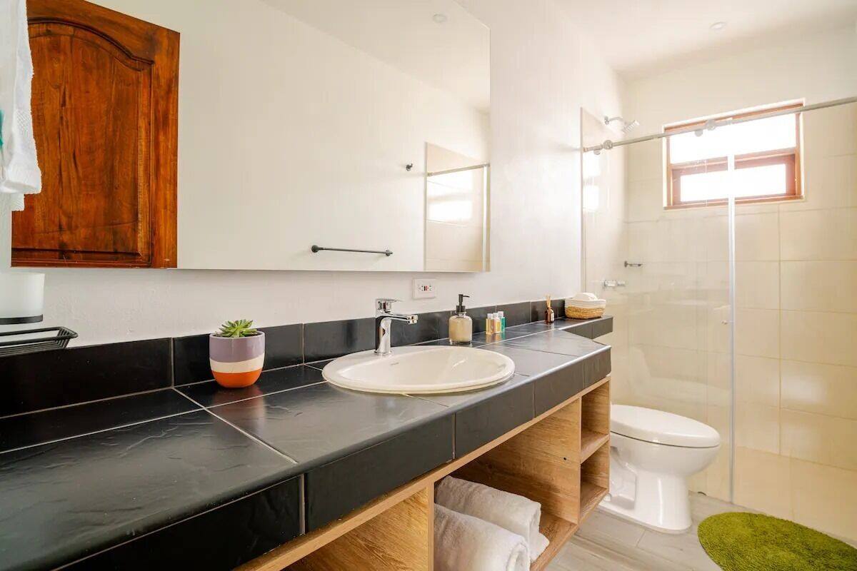 A modern bathroom with a black countertop, white sink, wooden vanity, and a glass shower enclosure.