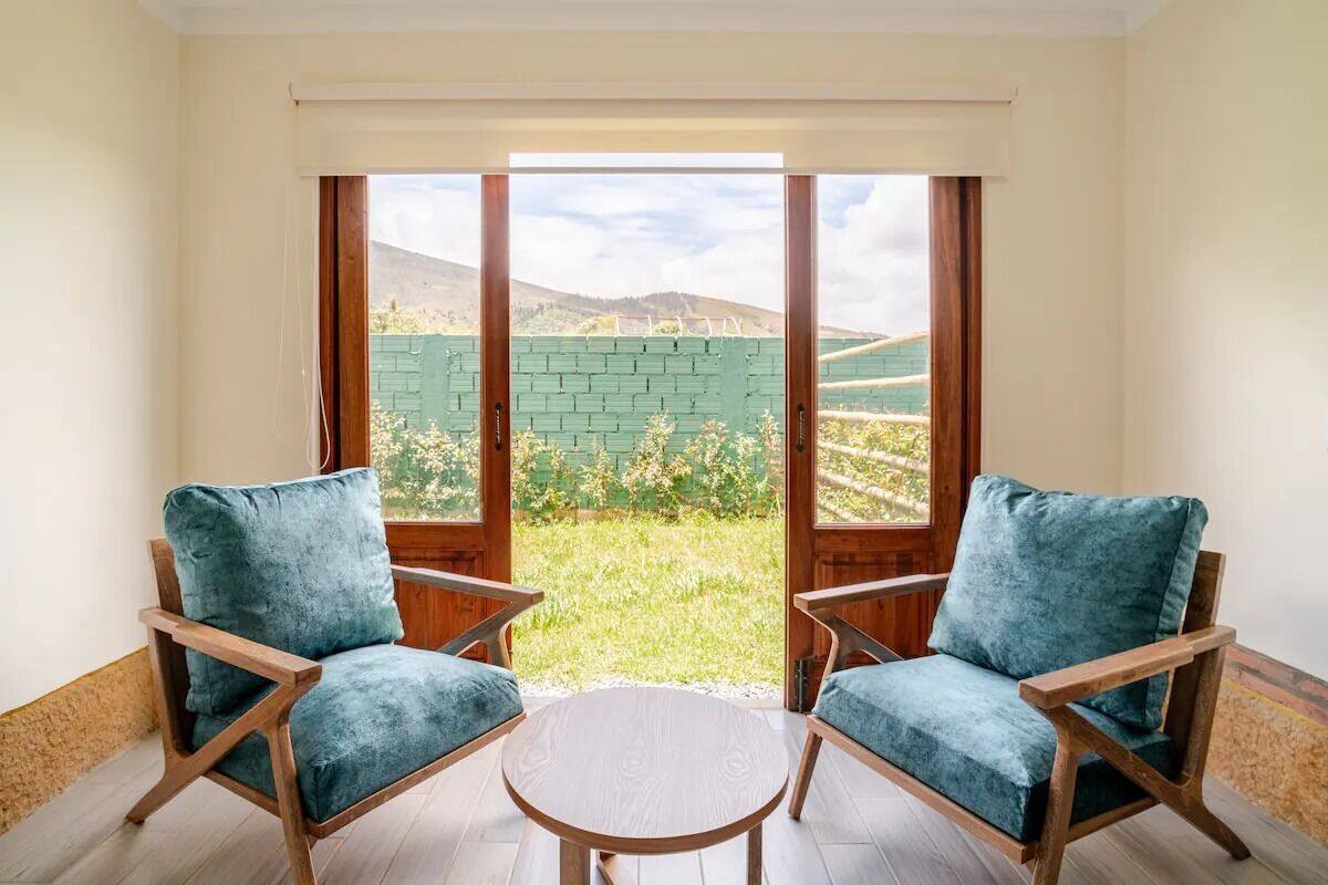 A room with two wooden chairs and a small round table, large windows with a view of a green field and hills.