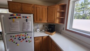 Una cocina con armarios de madera, un refrigerador blanco con imanes, una cafetera y una ventana con vistas a árboles.