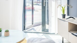 Una sala de estar moderna con un sofá, una mesa redonda con dos plantas en macetas, un televisor sobre un mueble bajo y un amplio ventanal con vistas al exterior.