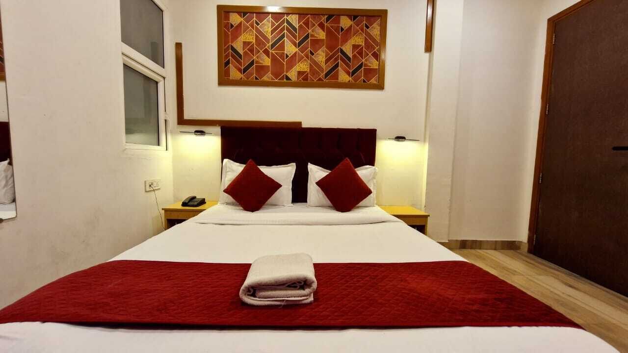 Een hotelkamer met een groot bed, een rode deken, twee rode kussens, een houten hoofdeinde, een nachtkastje met een telefoon en een kunstwerk boven het bed.