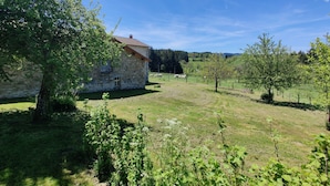 Une maison en pierre avec un toit en pente, entourée d’un champ herbeux et d’arbres.
