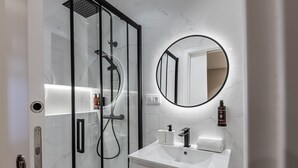 Casa de banho moderna com chuveiro em cabine de vidro, espelho redondo e lavatório branco com torneira preta.