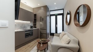 Cozinha moderna com bancada de mármore, forno embutido e frigorífico de aço inoxidável. Há uma mesa redonda de madeira com um copo e um tabuleiro. Um sofá bege com almofada macia está na área de estar, e dois espelhos de madeira estão na parede.