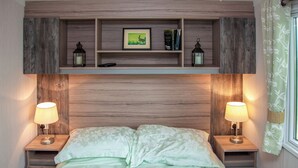 A bedroom with a wooden headboard, a bed with floral bedding, two bedside lamps, a shelf with books and a framed picture, and a window with a curtain. - 2 bedroom accommodation in West Quantoxhead, near Watchet (West Quantoxhead, near Watchet)