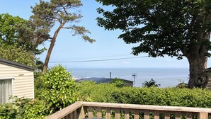 A wooden deck with a glass table and chairs, overlooking a body of water and a distant shoreline. - 2 bedroom accommodation in West Quantoxhead, near Watchet (West Quantoxhead, near Watchet)