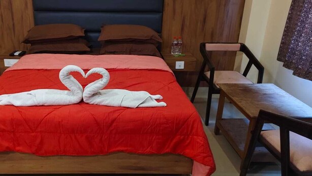 Une chambre d  ’hôtel avec une couvre-lit rouge, une tête de lit en bois, deux oreillers marron, une table de chevet en bois avec une bouteille, et une chaise en bois.