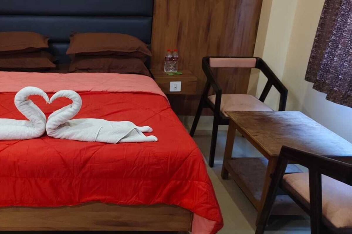 A hotel room with a red bedspread, a wooden headboard, two brown pillows, a wooden nightstand with a bottle, and a wooden chair.