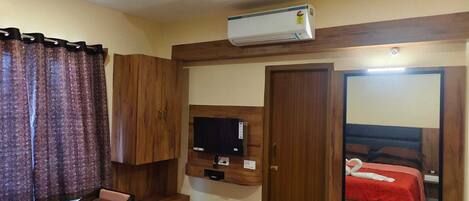 Una habitación de hotel con un armario de madera, un televisor montado, una cama con colcha roja y un equipo de aire acondicionado.