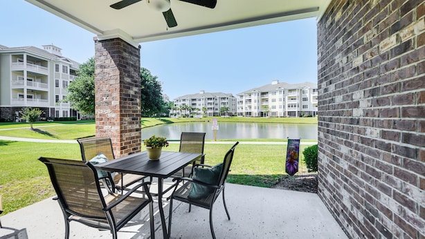 A covered patio with a table and chairs, a brick pillar, and a view of a lake and apartment buildings in the background.