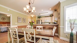 A dining area with a wooden table and white chairs, a chandelier, and a kitchen in the background.