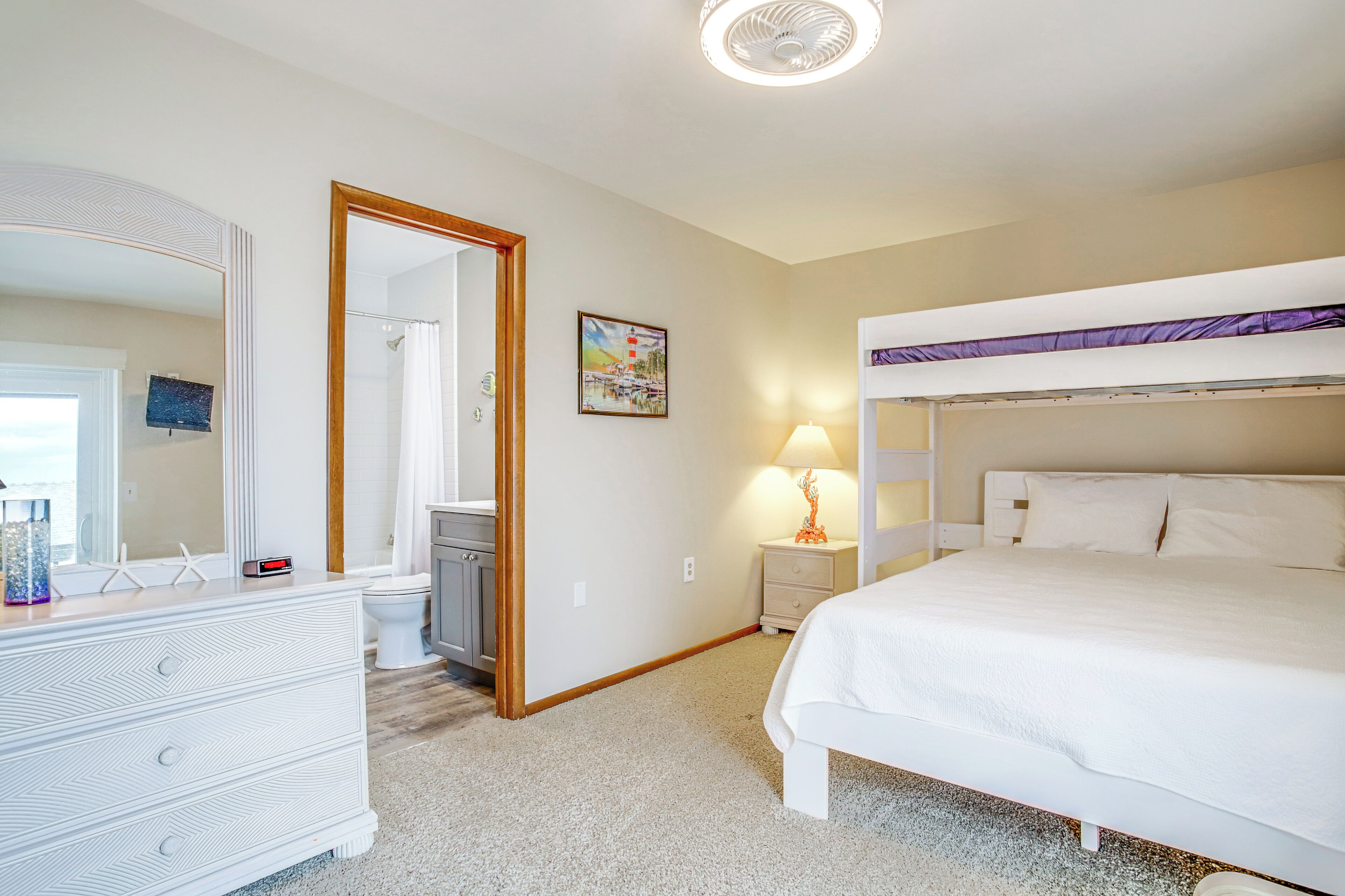 A bedroom with a bunk bed, a dresser, a mirror, a bedside table with a lamp, and a painting on the wall.