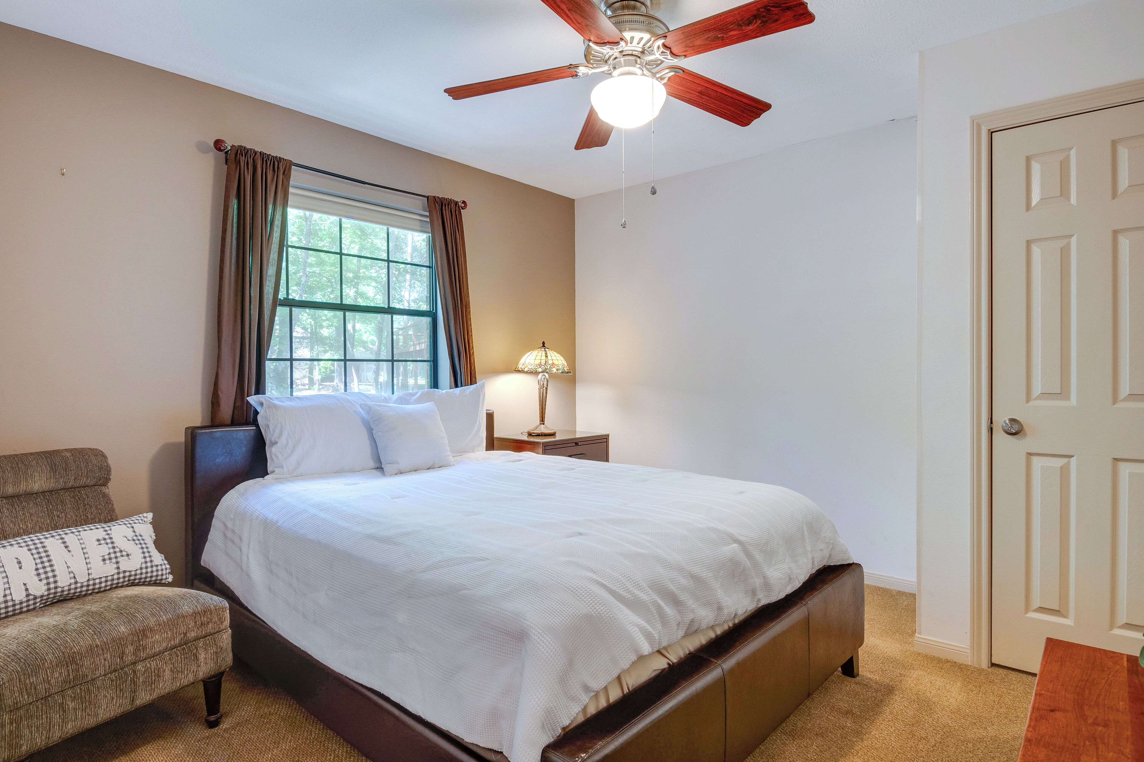 A bedroom with a bed, a chair, a nightstand, a lamp, a ceiling fan, and a door.