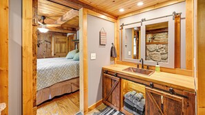 Interior rústico de cabaña con techo de madera, cama con colcha floral, banca de madera con toallas, lavamanos con salpicadero de piedra y espejo con pared de piedra de fondo.