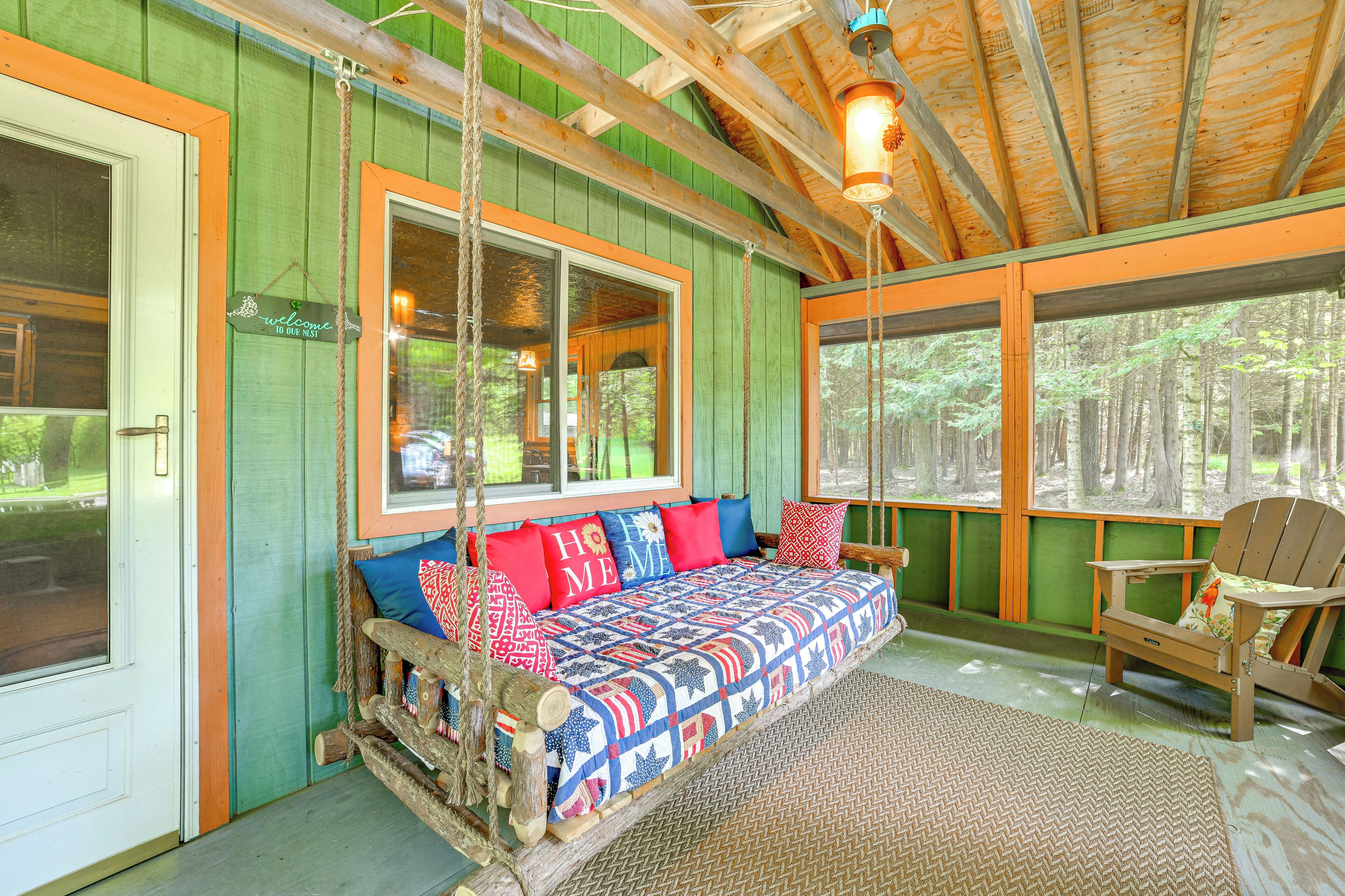 A cozy cabin interior with a wooden swing bed, a rocking chair, and a large window overlooking a forest.