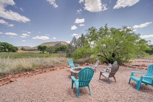 An outdoor seating area with four Adirondack chairs around a fire pit, set on a gravel surface with a view of a hilly landscape and trees under a clear sky.