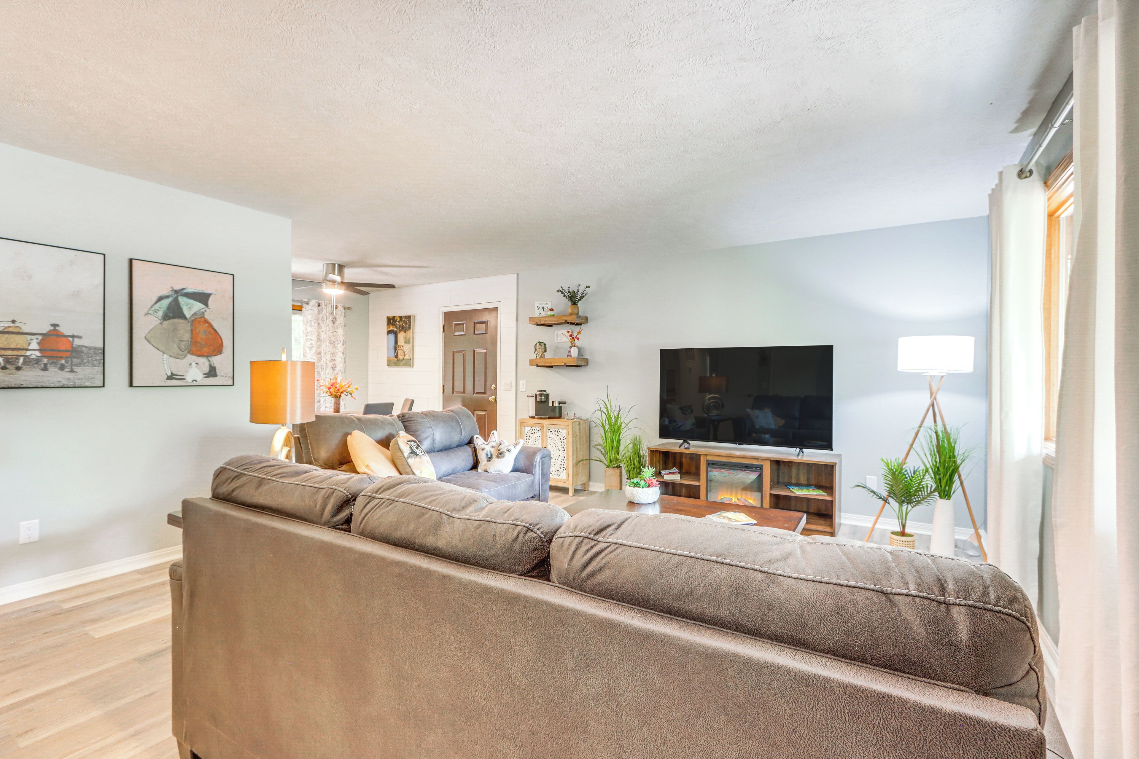 A living room with a brown leather sofa, a flat-screen TV, and a coffee table. There are decorative items on shelves and a potted plant on a side table.