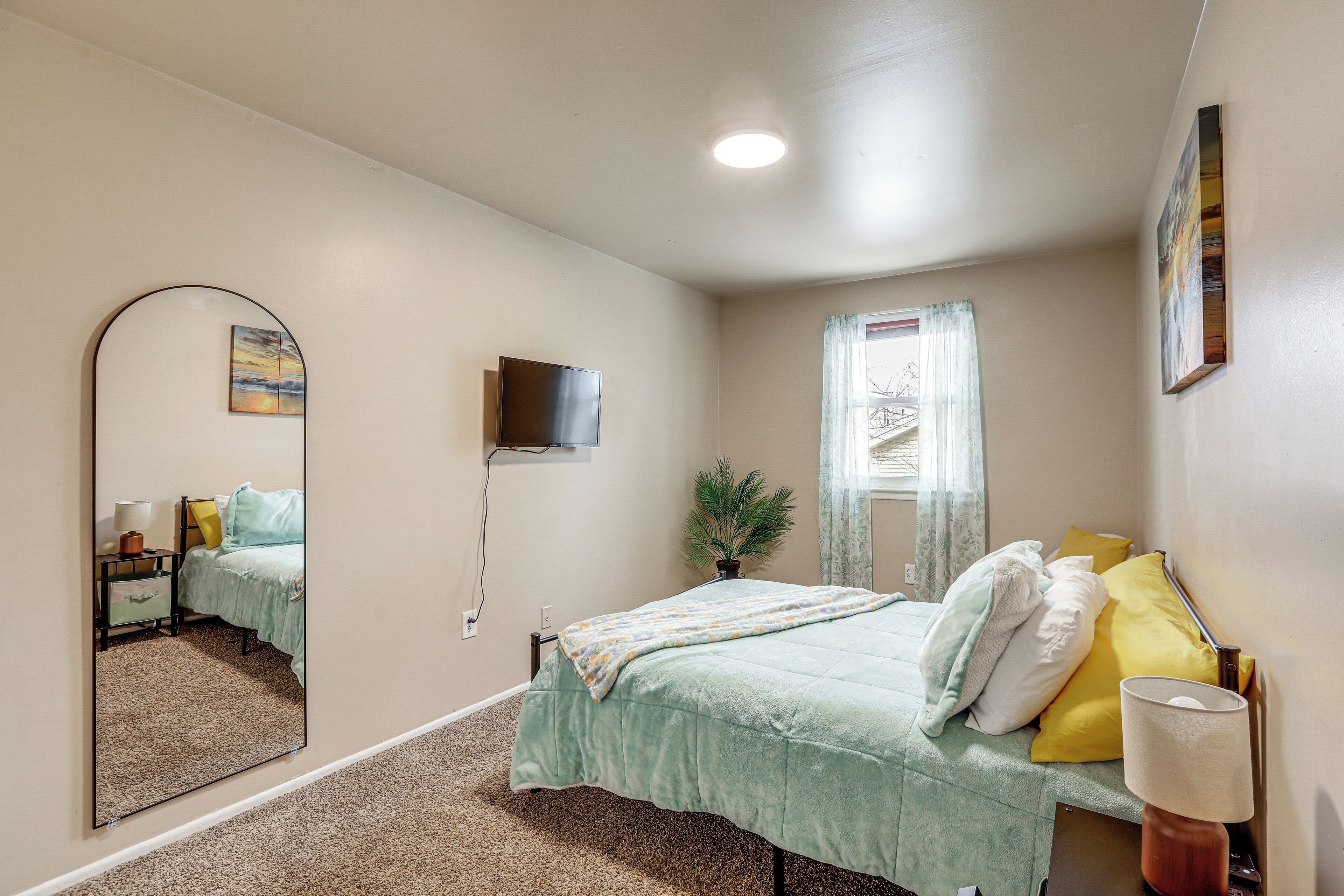 A bedroom with a bed, a mirror, a television, a window with curtains, and a plant.