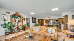 A spacious living room with a wooden coffee table, a leather sofa with cushions, a bookshelf, and a kitchen in the background. - Oro Valley Retreat w/ Pool & Breathtaking Views! (Oro Valley)