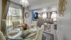 Ein Essbereich mit einem Glastisch, weißen Tellern und einer Chandelier. Im Hintergrund befindet sich eine Couch und ein dekoratives Wandelement.